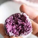 a woman's hand holding a half eaten blueberry energy ball covered in shredded coconut