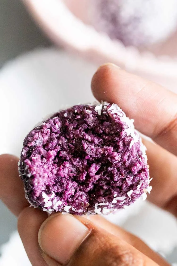 a woman's hand holding a half eaten blueberry energy ball covered in shredded coconut